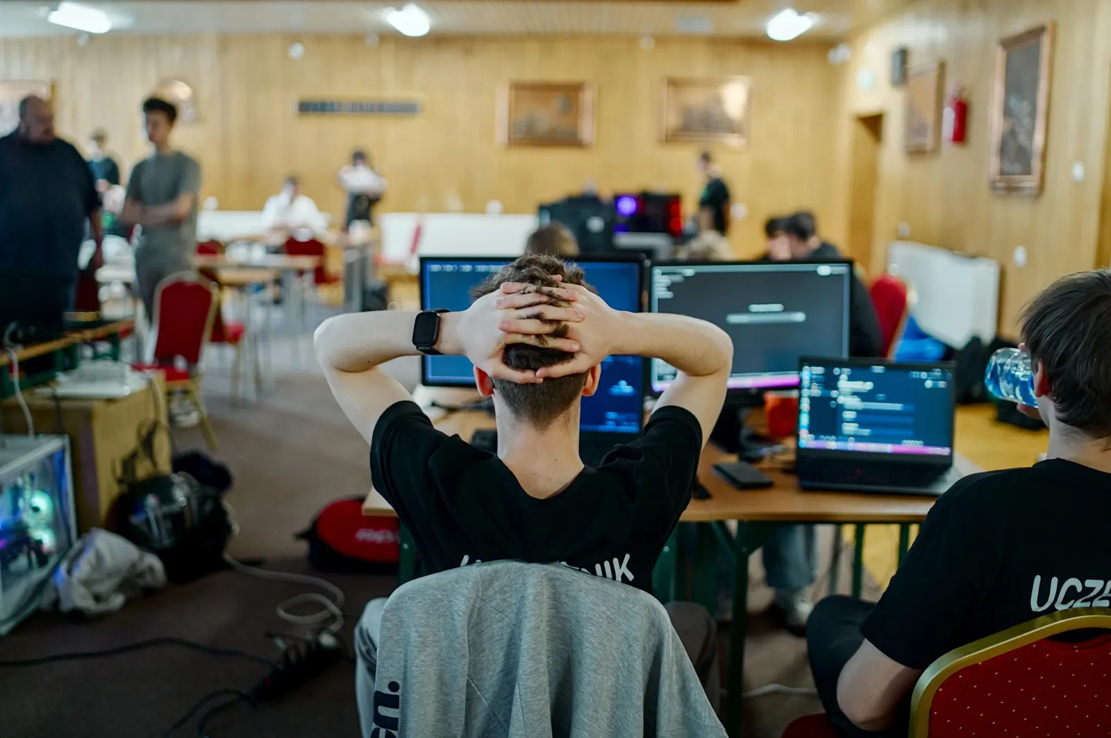 Uczestnik HackEDU z rękami za głową przed monitorami, w tle sala pełna zespołów